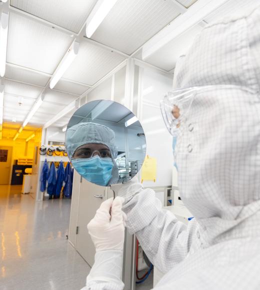 Cleanroom technician in white suit holds reflective wafer showing masked face