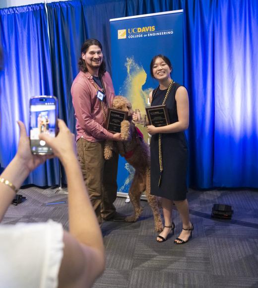 Woman photographing three smiling colleagues on blue-curtained stage, one holding plaque