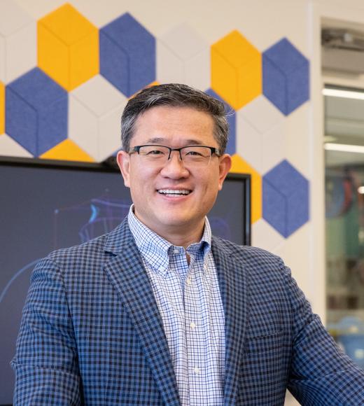 Smiling man with glasses and blazer in office; blue and yellow hexagonal wall panels